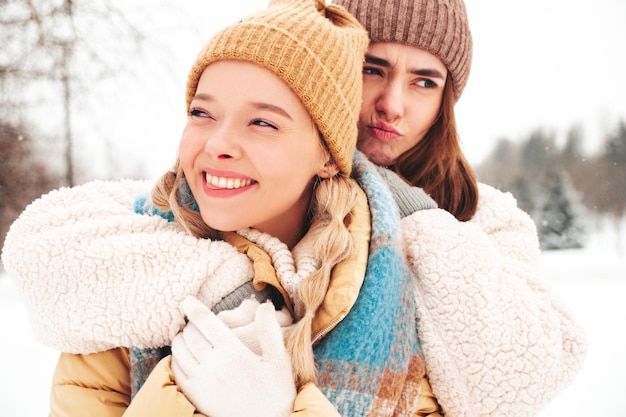 Due giovani belle donne sorridenti hipster in vestiti caldi alla moda e sciarpe. donne spensierate in posa in strada nel parco. modelli puri positivi che si divertono nella neve. godersi i momenti invernali. concetto di natale