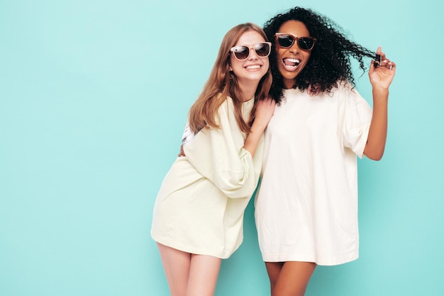 Two young beautiful smiling international hipster female in trendy summer clothes Sexy carefree women posing near blue wall in studio Positive models having fun in sunglasses Concept of friendship