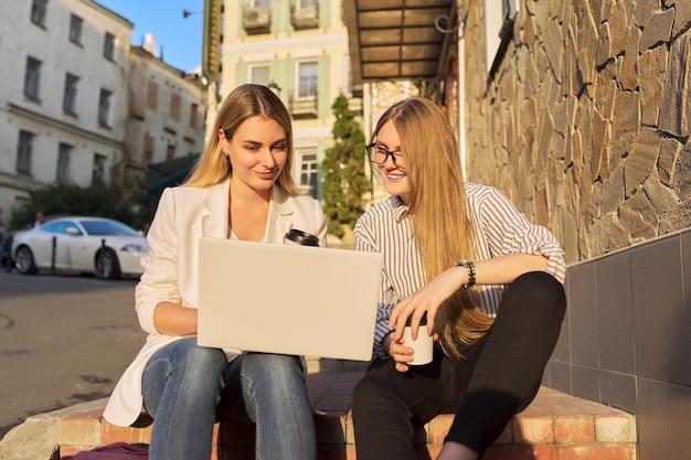 Two young beautiful smiling women looking at laptop monitor, sitting outdoors on the steps in city. Rest, friendship, leisure, lifestyle, technology