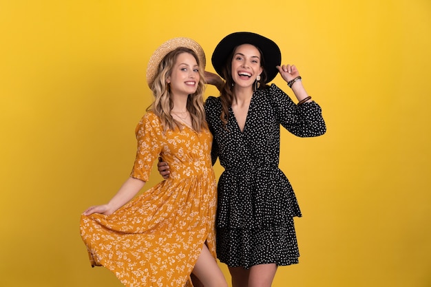 Two young beautiful women friends together isolated on yellow background in black and yellow dress and hat