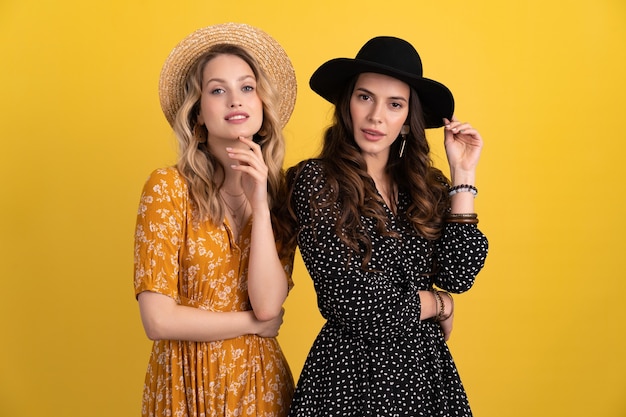 Two young beautiful women friends together isolated on yellow background in black and yellow dress and hat