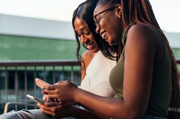 Two young black women laughing happily while consulting their mobile phones