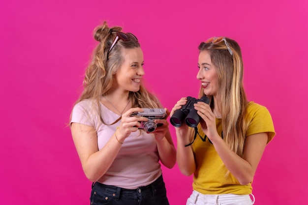 Two young blonde caucasian women enjoying vacation with a camera and spyglasses isolated on a pink background