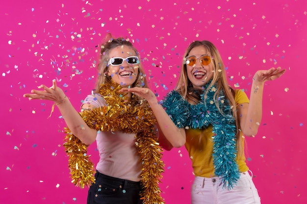 Two young blonde caucasian women partying at the nightclub having fun throwing confetti isolated on a pink background