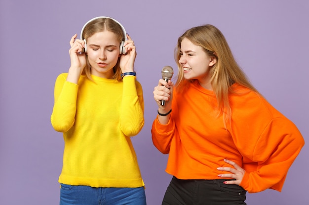 Foto due giovani sorelle gemelle bionde in abiti colorati ascoltano musica con le cuffie, cantano una canzone nel microfono isolato sulla parete blu viola. concetto di stile di vita familiare di persone.