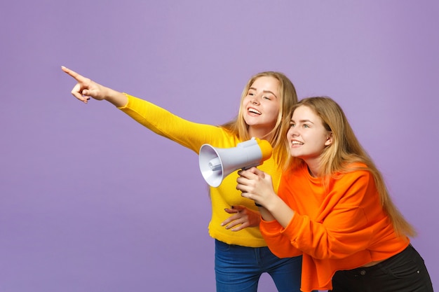 Two young blonde twins sisters girls in colorful clothes pointing index finger aside scream on megaphone isolated on pastel violet blue wall . People family lifestyle concept. 