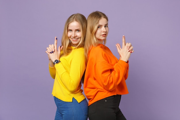 Two young blonde twins sisters girls in colorful clothes standing back to back, keeping fingers like gun isolated on pastel violet blue wall . People family lifestyle concept. 