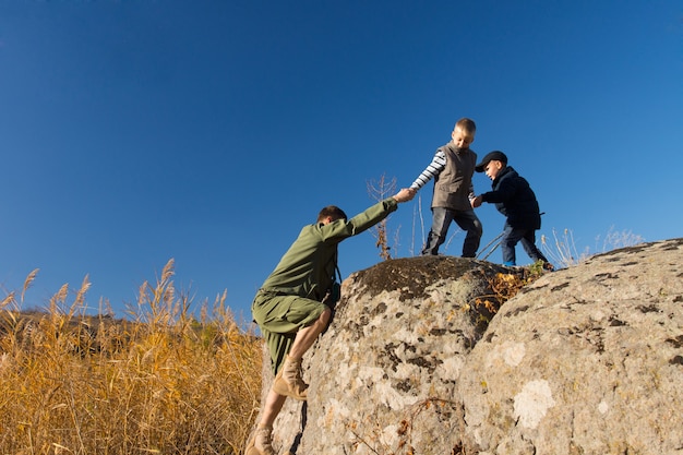 Two young boys helping a scout climb a rock cliff pulling his hand to assist him over the edge onto the summit