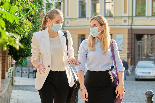 Two young business women in protective medical masks walking together along the city street. Lifestyle, business in an epidemic, pandemic, bad ecology