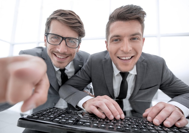 Two young businessman are looking into the camera while sitting in the office One shows the index finger