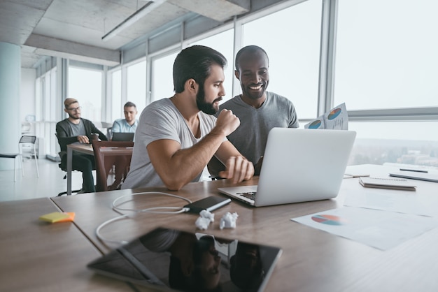 Two young businessmen using laptop and discussing new project at office, developing strategy for online business, explaining sharing ideas, preparing presentation, having brainstorming session