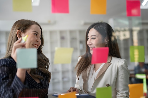 두 명의 젊은 여성 사업가 크리에이티브 팀이 유리 벽에 포스트잇 노트를 사용하여 개발 전략 사업 계획을 작성하여 성공으로 성장합니다.
