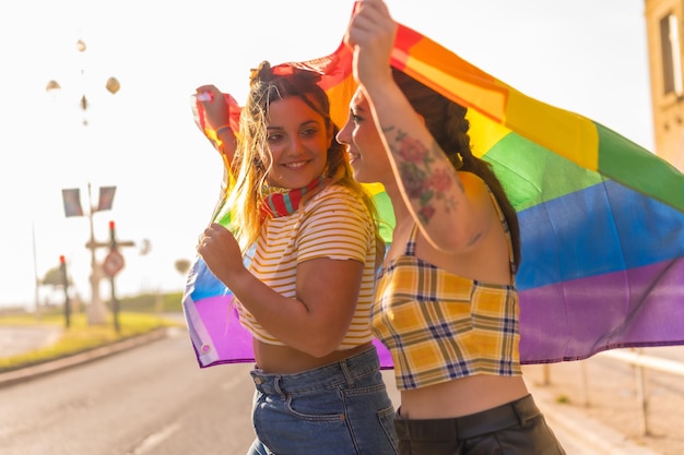 Due giovani femmine caucasiche che abbracciano con la bandiera dell'orgoglio lgbt all'aperto