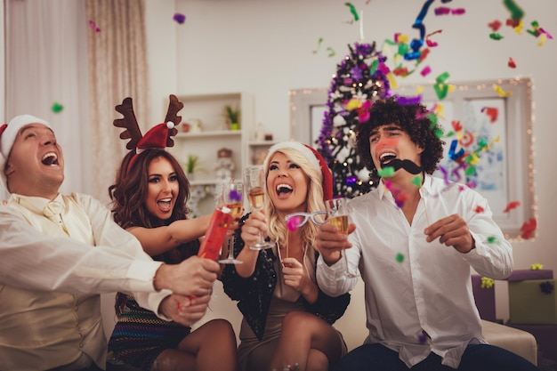 Две молодые веселые пары празднуют Рождество с конфетти дома. Они веселятся и пьют шампанское.