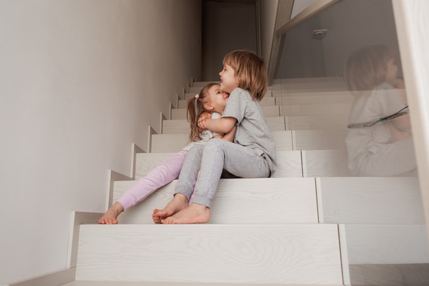 Двое маленьких детей, брат и сестра, обнимаются дома на деревянной лестнице. Фото высокого качества