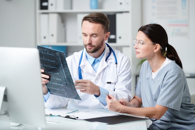Два молодых врача в униформе сидят за столом перед монитором компьютера, обсуждают рентгеновский снимок пациента и консультируются в медицинском кабинете