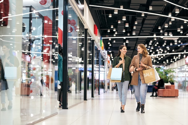 Two young contemporary females in elegant casualwear walking along shopwindows inside mall and discussing what they would like to buy