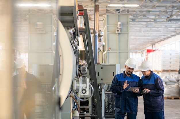 Фото Двое молодых современных рабочих современного завода обсуждают онлайн технические данные или эскизы деталей для промышленных машин в мастерской