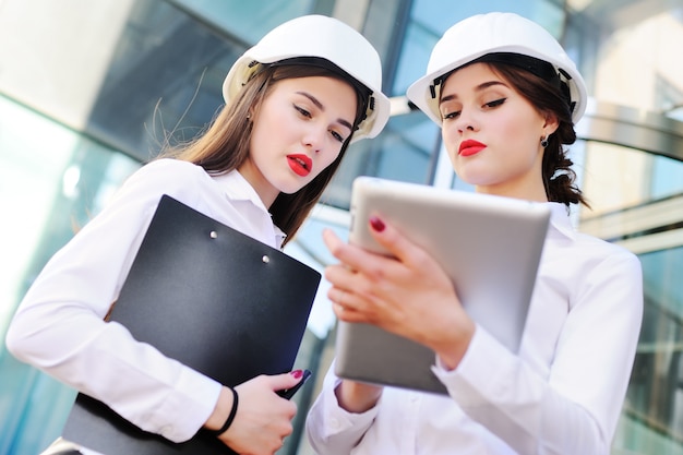 Two young cute girls in business clothes and white construction helmets discuss a business plan or contract