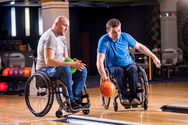 Двое молодых инвалидов в инвалидных колясках играют в боулинг в клубе