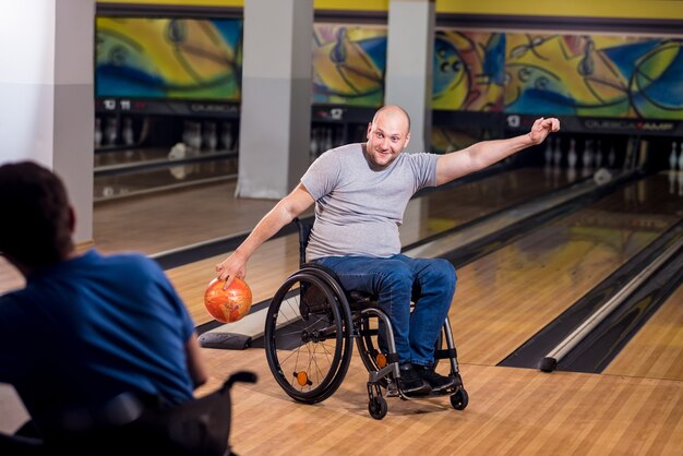 Двое молодых инвалидов в инвалидных колясках играют в боулинг в клубе