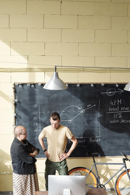 コンピューターモニターの前で黒板のそばに立って、会議でオンラインデータについて話し合う2人の若いエコノミスト