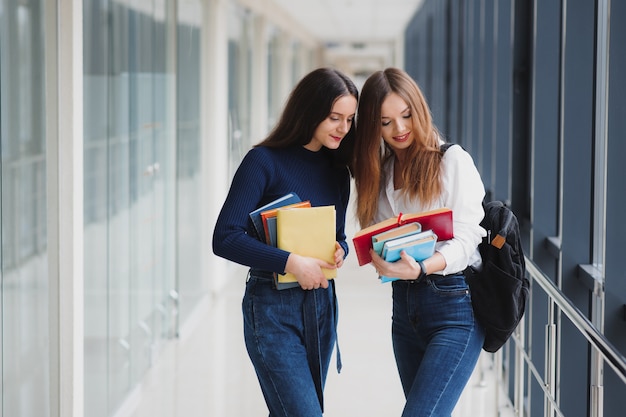 Two young female students standing with books and bags in the hallway