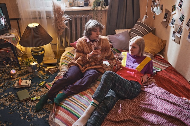 Two young friends lying on bed talking to each other and drinking red wine in the room