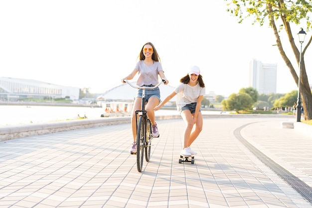 Two young friends relaxing on a hot summer day with one young woman riding a bicycle towing her friend on a skateboard down an urban street.