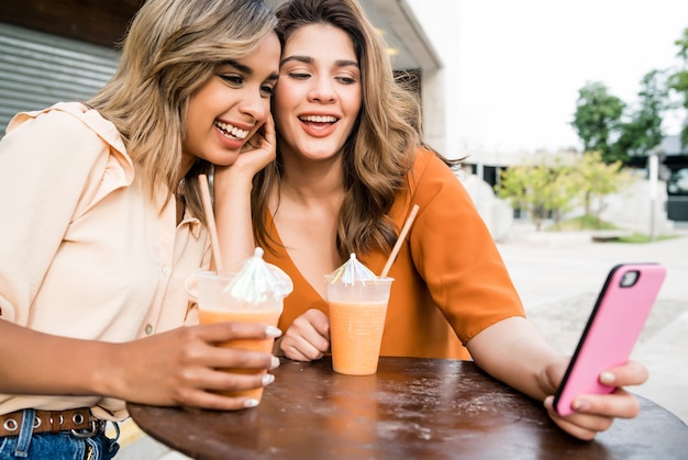Двое молодых друзей используют свой мобильный телефон и пьют свежий фруктовый сок в кафе на открытом воздухе