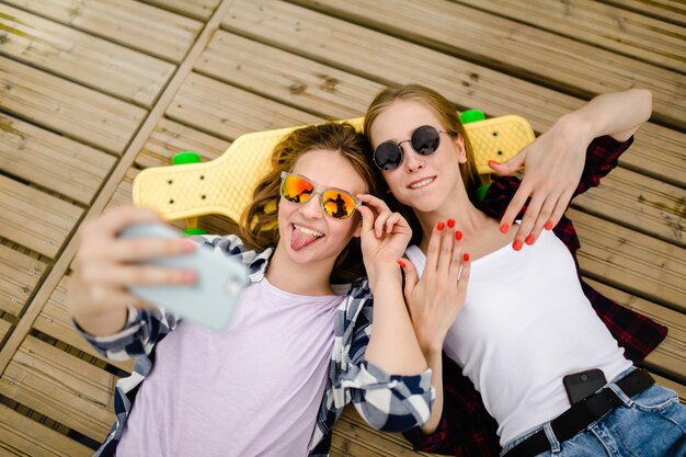 Two young girl in hipster outfit making selfie while lying with on wooden pier.