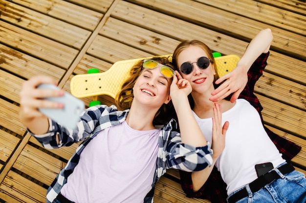 Photo two young girl in hipster outfit making selfie while lying with on wooden pier.