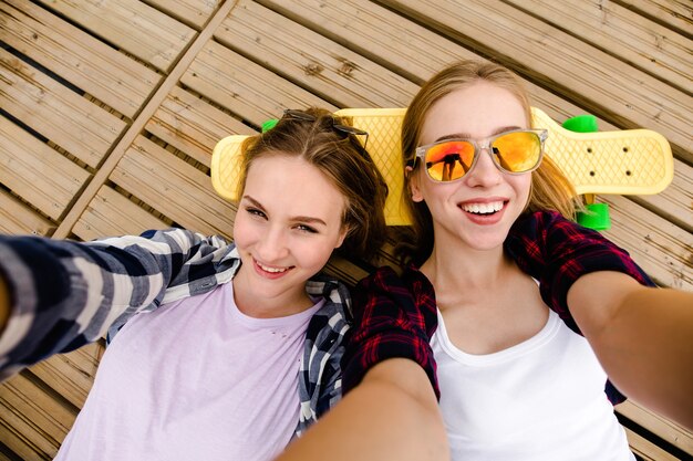 Two young girl in hipster outfit making selfie while lying with on wooden pier.