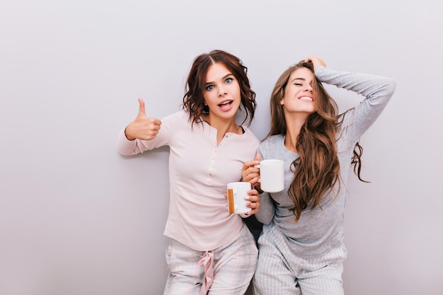 Two young girls in pajamas having fun on gray wall background. Girl with long hair laughing and keeps eyes closed, other with curly hair smiling to camera