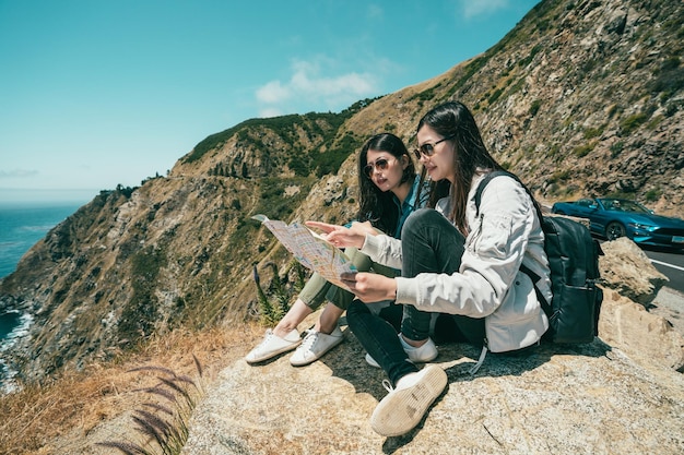 Two young girls reading the guide map and sitting on the cliff\
while a beak time with a background of stunning mountains and\
sea.