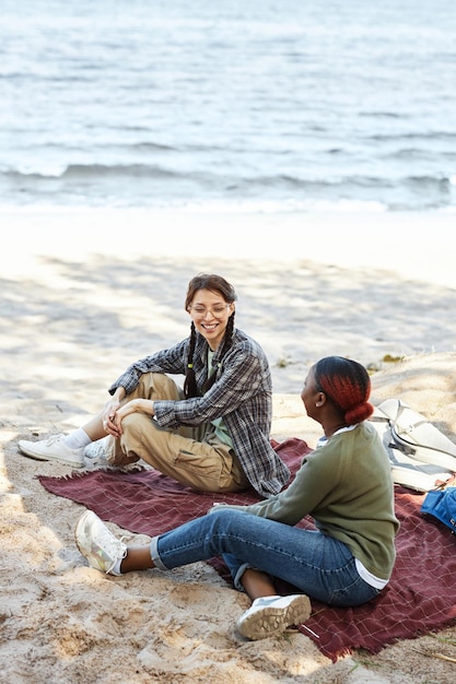 Two young girls sitting on seashore and talking to each other during picnic outdoors