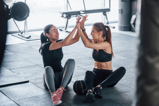 Photo two young girls in sportive clothes is in the gym together at daytime sitting on the floor with ball and giving high five.