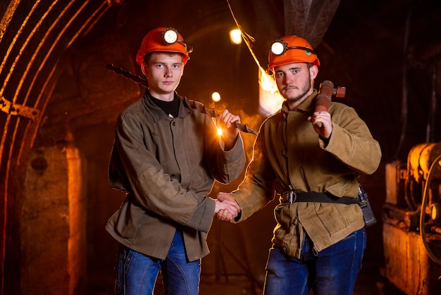 Photo two young guys in working uniform and protective helmets, shaking hands. workers of the mine. miners