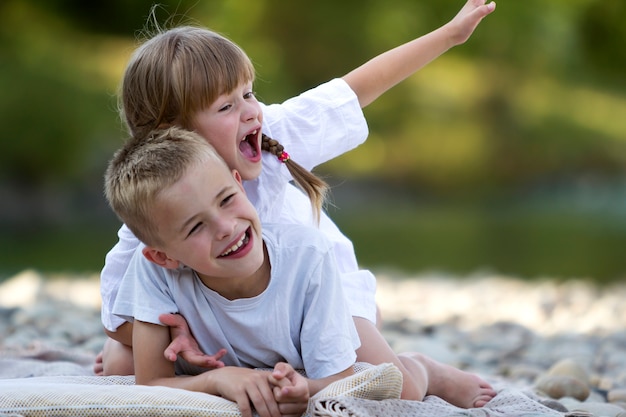 Two young happy cute blond laughing children, boy and girl, brother and sister having fun on pebbled beach on blurred bright sunny summer bokeh . Friendship and perfect holiday concept.