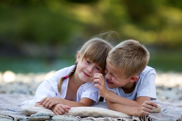 Two young happy cute blond smiling children, boy and girl, brother and sister laying embraced on pebbled beach on blurred bright sunny summer day background. Friendship and perfect holiday concept.