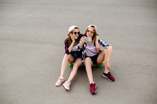 流行に敏感な衣装を着た2人の若い幸せなガールフレンドがロングボードに一緒に座って、電話で自分撮りをしています。