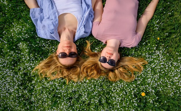 Two young happy girls with long hair lie on green grass on sunny summer day and smile at camera