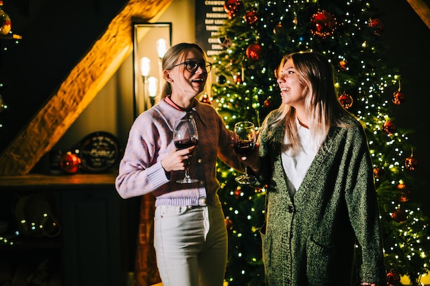 Two young happy women friends having fun, drinking wine near Christmas tree, celebrating winter holidays