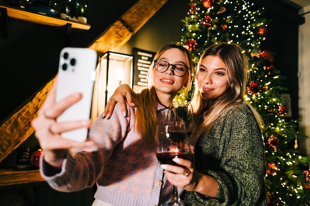 写真 クリスマスツリーの近くでお祝いの写真selfieを作り、ワインを飲む2人の若い幸せな女性。