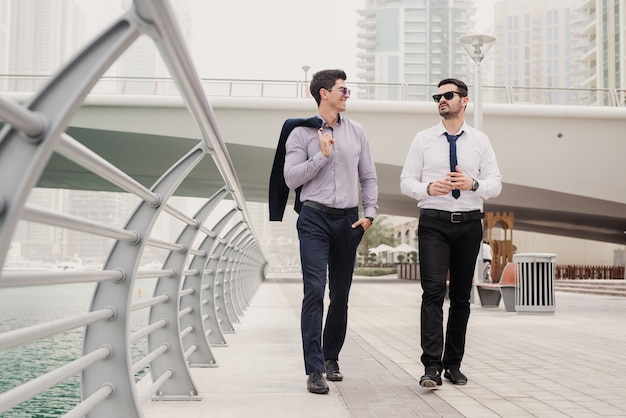 Two young hardworking businessman in suit walking through Dubai Marine and talking about business situation in their office.