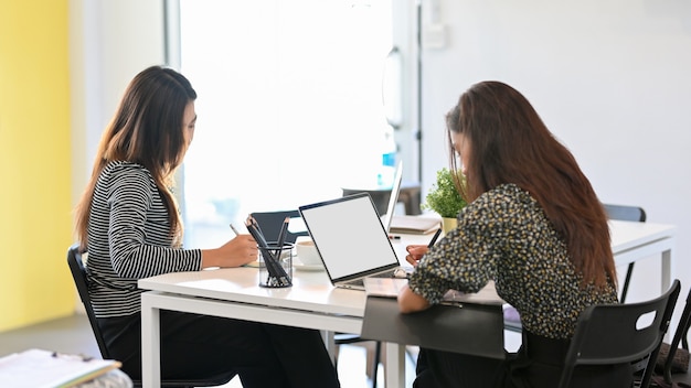 Две молодые трудолюбивые женщины, работающие в офисе с электронными устройствами, с помощью ноутбука