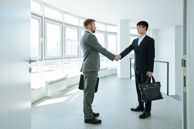 Two young intercultural business partners in formalwear standing in front of each other and handshaking in corridor inside office center