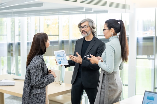 Two young intercultural businesswomen listening to mature business partner with digital tablet presenting financial charts and diagrams