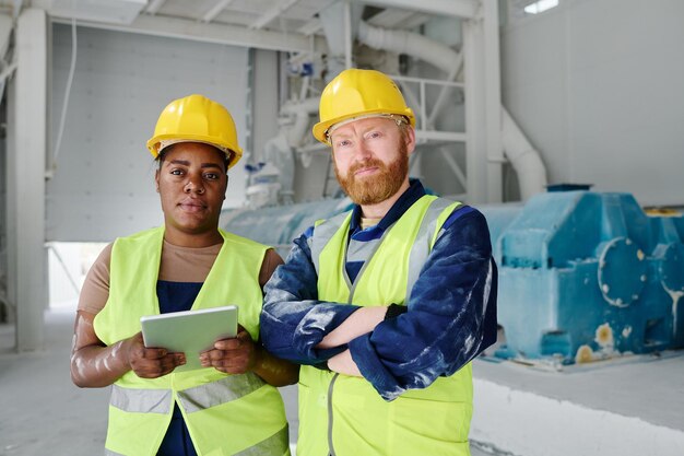 Two young intercultural colleagues in uniform and hardhats looking at you