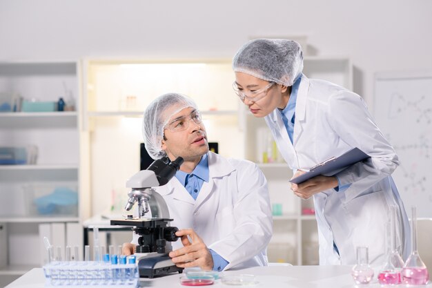 Two young intercultural scientists in whitecoats consulting in lab while working over new vaccine and studying viruses